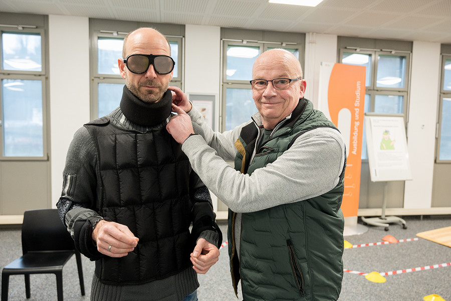 Ein Mitarbeitender testet den GERT-Alterssimulationsanzug. Ein Kollege hilft beim Ankleiden.