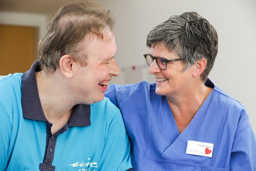 Viele Menschen mit Behinderung haben dringenden Bedarf an einer Begleitung im Krankenhaus. Auf dem Bild sitzen ein erkrankter Mann und seine Betreuerin nebeneinander und lächeln sich an.