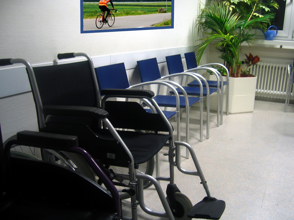 Das Bild zeigt einen Rollstuhl neben normalen Stühlen in einem Wartezimmer.