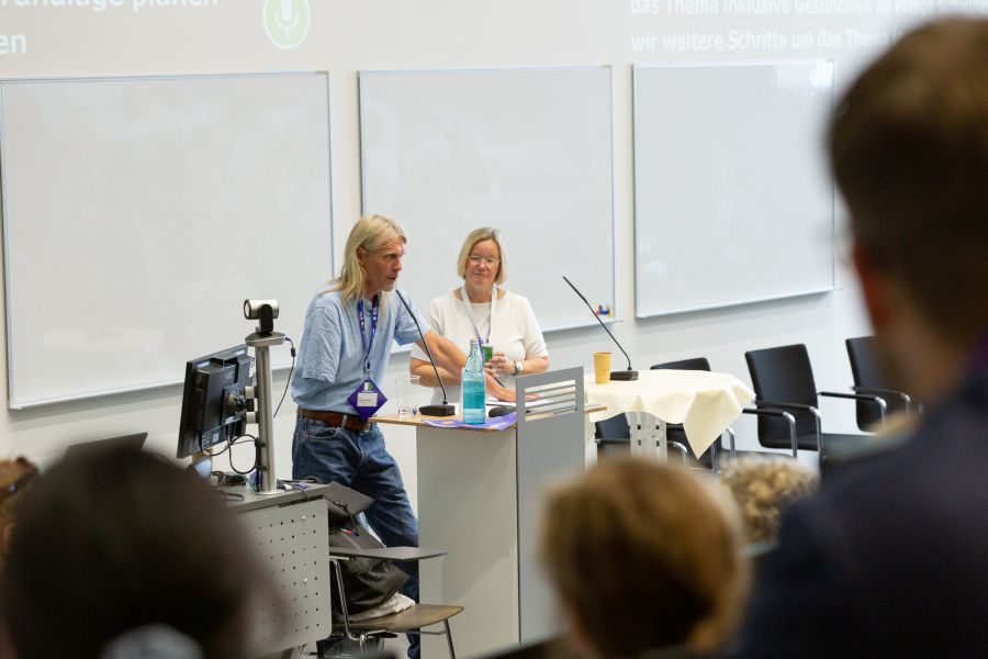 Ein mann mit längeren Blonden Haaren spricht an einem Pult vor Publikum in einem Hörsaal ähnlichem Ort