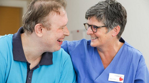 Viele Menschen mit Behinderung haben dringenden Bedarf an einer Begleitung im Krankenhaus. Auf dem Bild sitzen ein erkrankter Mann und seine Betreuerin nebeneinander und lächeln sich an.