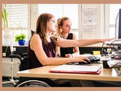 Zwei Frauen sitzen an einem Schreibtisch. Sie schauen gemeinsam auf einen Bildschirm. EDie Frau im Vordergrund sitz im Rollstuhl. Sie zeigt mit dem Zeigefinger der linken Hand auf den Bildschirm.