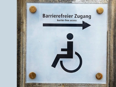 Das Foto zeigt das Symbol für einen Menschen im Rollstuhl. Ein Pfeil weist nach links. Darüber steht der Schriftzug Barrierefreier Zugang.