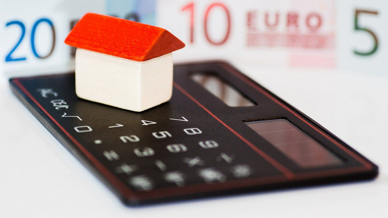 Ein kleines Holzhaus steht auf einem Taschenrechner. Im Hintergrund EURO-Geldscheine.