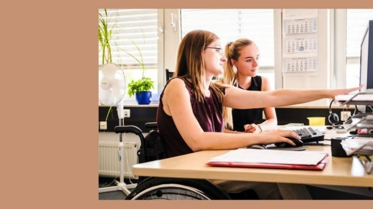 Zwei junge Frauen sitzen gemeinsam am Schreibtisch und betrachten einen Computer-Monitor. Eine Frau sitzt in einem Rollstuhl.