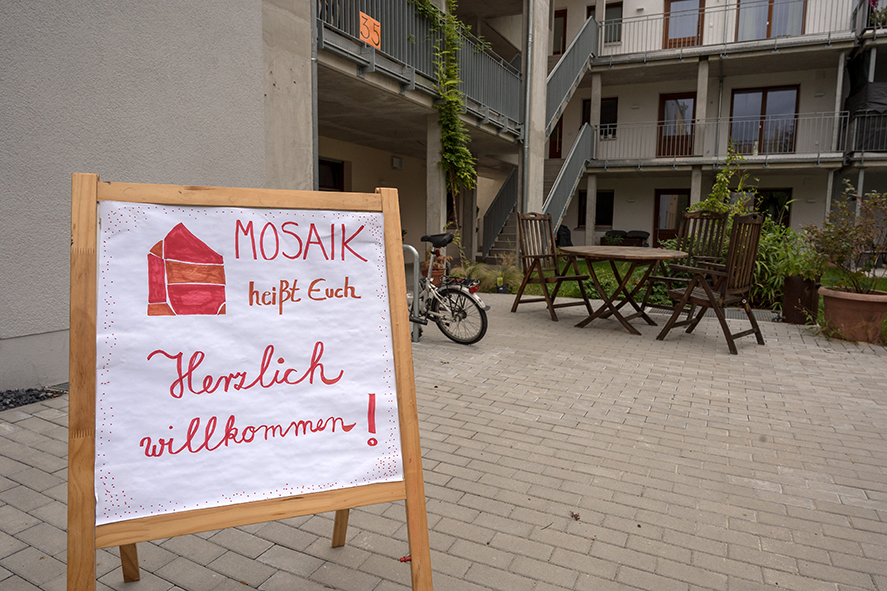 Das Foto zeigt einen Hof mit einen Willkommenschild: MOSAIK heißt Euch herzlich willkommen! steht darauf