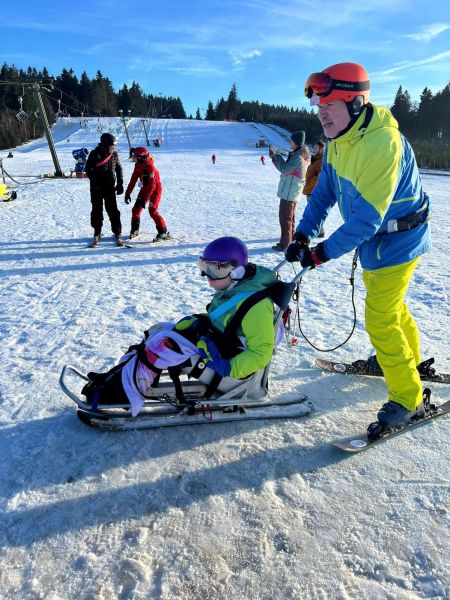 Ein Kind, das gut befestigt auf einem Skibob sitzt, wird von einem Erwachsenen auf Skiern über die Piste geschoben
