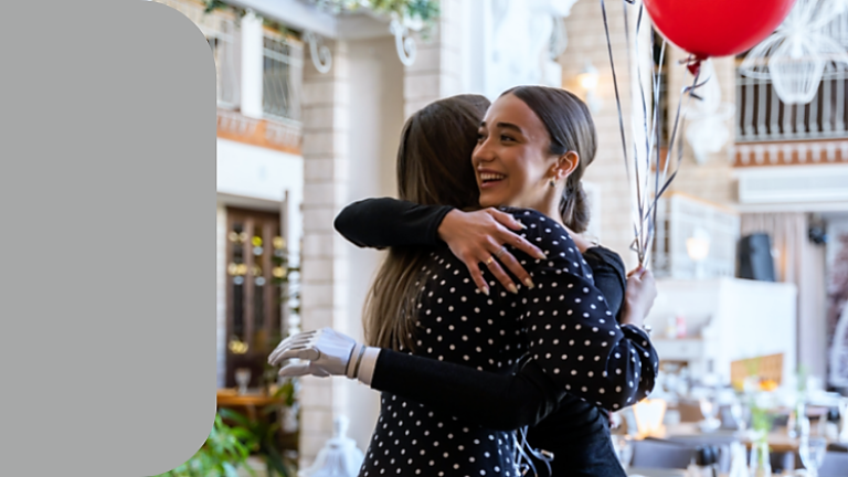 Zwei Frauen umarmen sich. Eine Frau hält einen roten Luftballon.